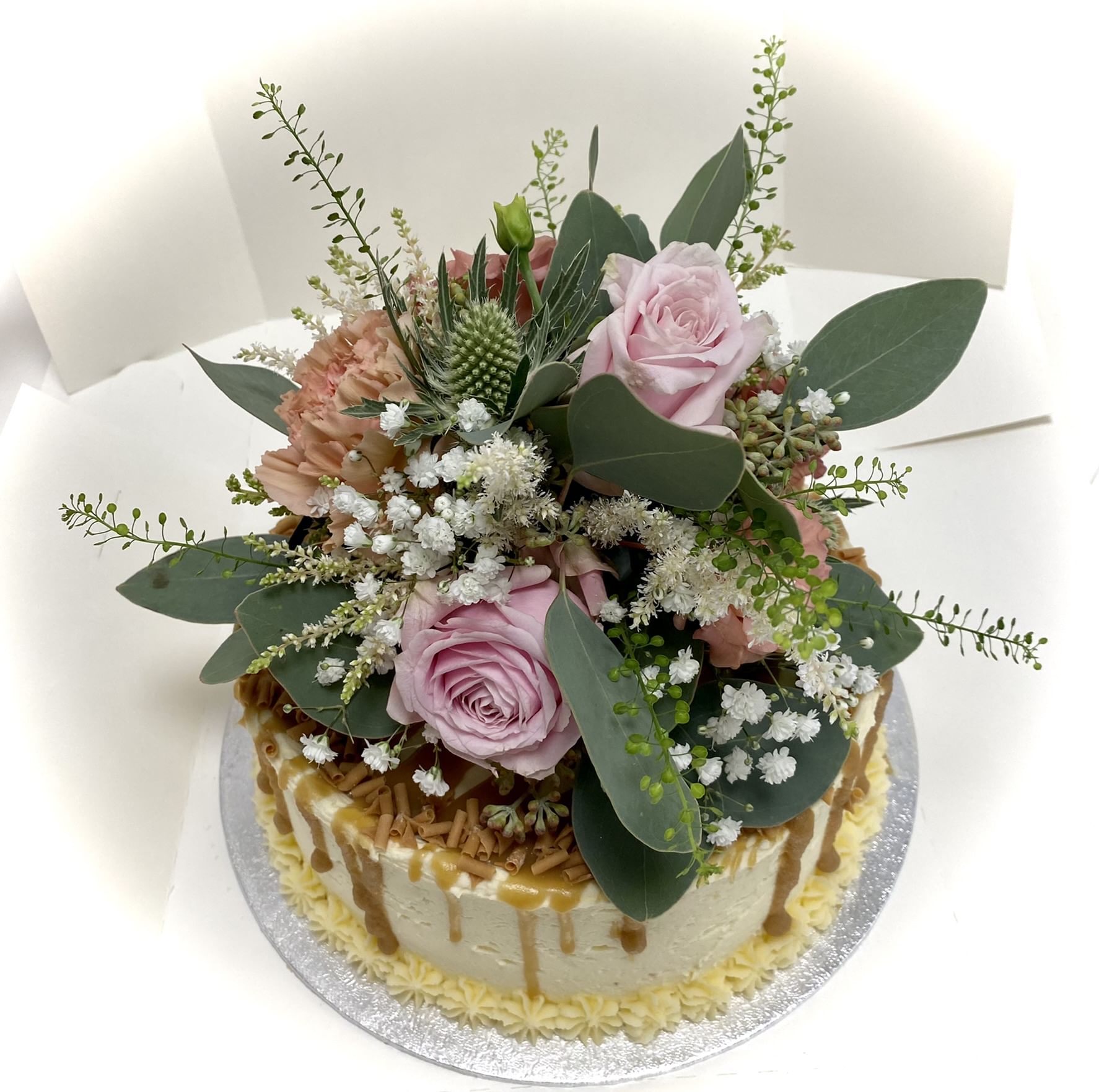 A Salted Caramel Cake adorned with an elegant arrangement of pink roses, baby’s breath, and lush greenery on top. The cake features decorative dollops of homemade salted caramel sauce cascading down the sides, with piped yellow flowers at the base.
